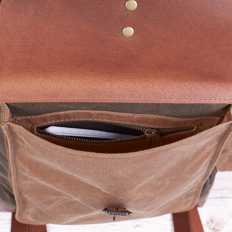 Puede incluir: Una solapa de cuero marr&oacute;n con un bolsillo con cremallera debajo. El bolsillo est&aacute; hecho de un material de lona marr&oacute;n y tiene un tirador de cremallera con un anillo de metal. La solapa se cierra con dos broches de presi&oacute;n de lat&oacute;n.