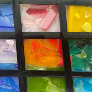 May include: A mosaic artwork made of small pieces of glass in various colors, including blue, green, yellow, orange, and pink. The glass pieces are arranged in a grid pattern, creating a geometric design.