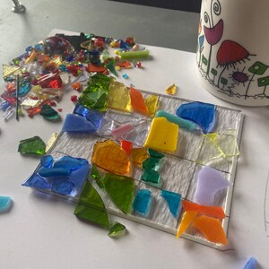 May include: A close-up of a mosaic art project in progress. The project is made of small pieces of glass in various colors, including blue, green, yellow, orange, and purple. The glass pieces are arranged on a white surface with a grid pattern.