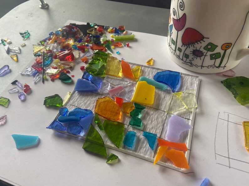 May include: A close-up of a mosaic art project in progress. The project is made of small pieces of glass in various colors, including blue, green, yellow, orange, and purple. The glass pieces are arranged on a white surface with a grid pattern.