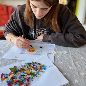 May include: A person is working on a mosaic art project. They are using tweezers to place small pieces of colorful glass onto a white surface. There are additional pieces of glass in a pile on a white sheet of paper.