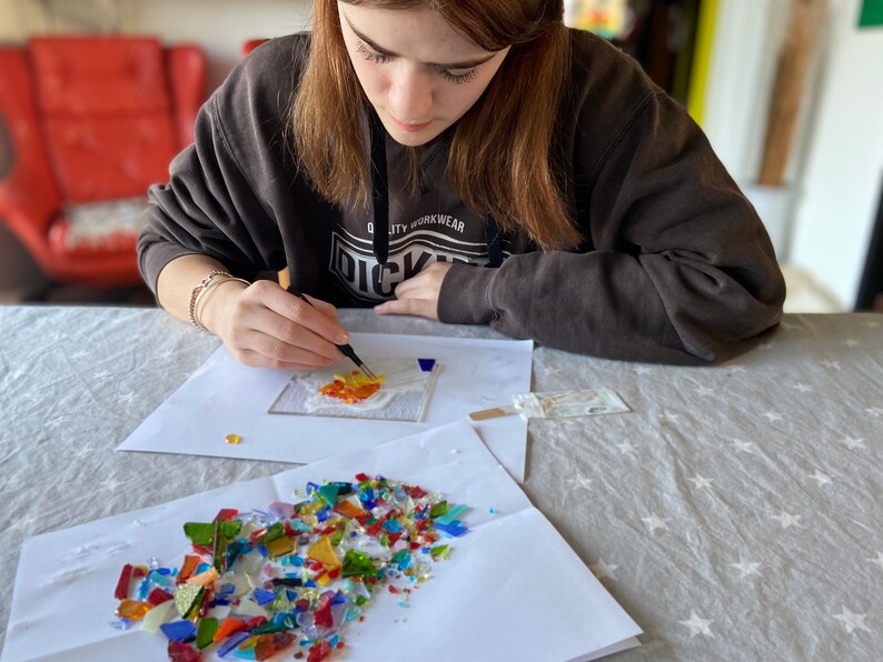 May include: A person is working on a mosaic art project. They are using tweezers to place small pieces of colorful glass onto a white surface. There are additional pieces of glass in a pile on a white sheet of paper.