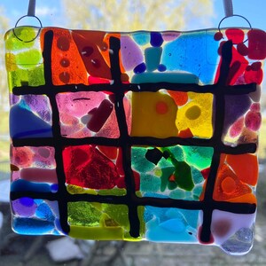 May include: A colorful stained glass window hanging with a grid pattern. The glass is made up of small pieces of different colors, including red, orange, yellow, green, blue, and purple. The grid lines are black.
