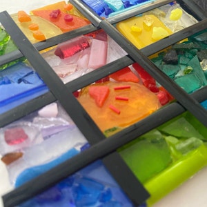 May include: A colorful mosaic artwork with various shades of blue, green, red, yellow, and orange glass pieces. The glass pieces are arranged in a grid pattern and framed with black borders.