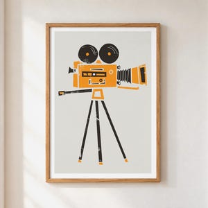 May include: A framed wall art print featuring a stylized graphic illustration of a vintage movie camera in orange and black on a light gray background. The camera is mounted on a black tripod, showcasing a retro design with two circular reels.