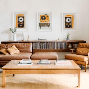 Puede incluir: Una sala de estar con un sof&aacute; de cuero marr&oacute;n, un sill&oacute;n de cuero marr&oacute;n y una mesa de caf&eacute; de madera. La mesa de caf&eacute; tiene tres pilas de libros encima. Hay tres impresiones enmarcadas en la pared sobre el sof&aacute;. Las impresiones son de un tocadiscos y altavoces vintage.