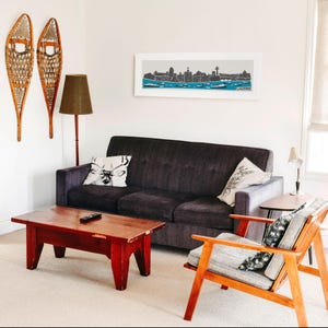 Puede incluir: Un interior de sala de estar con un sof&aacute; gris oscuro, una mesa de caf&eacute; de madera, un sill&oacute;n de madera y una impresi&oacute;n enmarcada de un horizonte de ciudad sobre el sof&aacute;. La impresi&oacute;n presenta una silueta en blanco y negro de la ciudad con la palabra "Chicago" en la parte inferior.