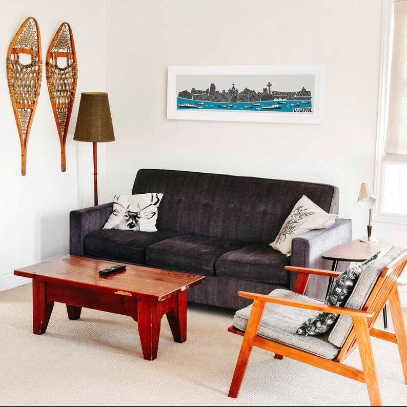 Puede incluir: Un interior de sala de estar con un sof&aacute; gris oscuro, una mesa de caf&eacute; de madera, un sill&oacute;n de madera y una impresi&oacute;n enmarcada de un horizonte de ciudad sobre el sof&aacute;. La impresi&oacute;n presenta una silueta en blanco y negro de la ciudad con la palabra "Chicago" en la parte inferior.
