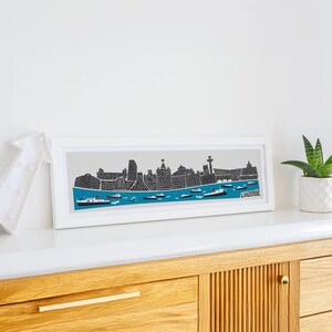 Puede incluir: Una impresi&oacute;n enmarcada en blanco de una ilustraci&oacute;n en blanco y negro del horizonte de Liverpool con una escena de agua azul y la palabra "Liverpool" en la esquina inferior derecha.