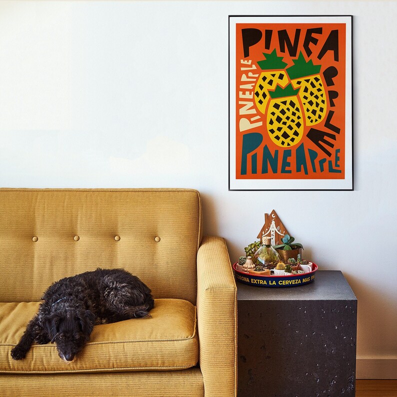 May include: A yellow couch with a black dog sleeping on it. A framed poster with the word "Pineapple" written in a colorful, stylized font is hanging on the wall above the couch. The poster features a graphic of three yellow pineapples.