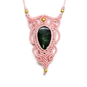 May include: A handmade macrame necklace featuring a teardrop-shaped, dark green stone pendant. The pendant is encased in a delicate, light pink macrame design with gold-colored beads. The necklace is suspended from a matching pink cord.