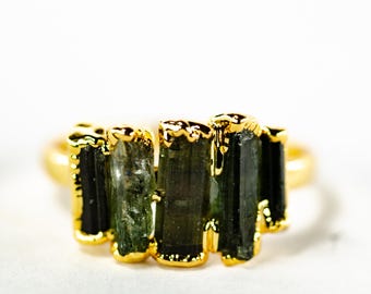 May include: A gold-colored ring featuring a cluster of dark green tourmaline crystals. The crystals are set in a gold-colored bezel setting, creating a unique and natural design. The ring is set against a white background.