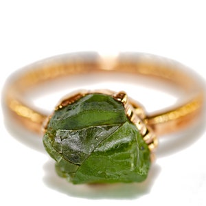 May include: A gold ring featuring a large, green, rough-cut gemstone. The gemstone is set in a gold bezel setting.