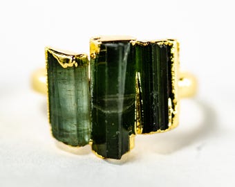 May include: A gold-colored ring featuring three rectangular, dark green tourmaline stones. The stones are set in a gold bezel setting, creating a striking contrast. The ring is displayed against a white background, highlighting the gemstone's natural beauty.
