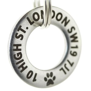 Puede incluir: Una placa de identificación de perro de plata con una huella de pata y el texto "10 HIGH ST. LONDON SW19 7JL" grabado en ella.