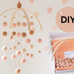 May include: DIY baby mobile kit with wooden hoops, pink, beige, and white felt balls, and instructions. The kit is in a white box with a peach-colored fabric lining.