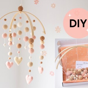 May include: A handmade baby mobile with a wooden frame, featuring hanging felt balls and heart shapes in shades of pink, beige, and brown. The DIY kit includes materials and instructions. The word "DIY" is displayed in a pink circle.