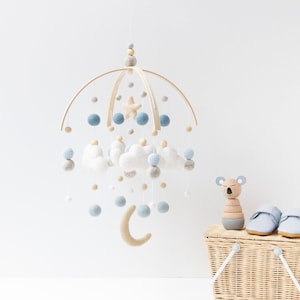 May include: A wooden baby mobile with a crescent moon, clouds, and stars. The mobile is made of felt and wood and is hanging from a wooden ring. The mobile is blue, white, and beige.