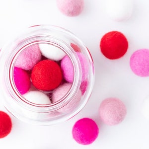 May include: A glass jar filled with pink, red, and white felt balls. The jar is sitting on a white surface with more felt balls scattered around it.
