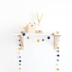 May include: A white shelf with a wooden bear figurine, a star, and two pencils. A garland of blue, white, and yellow felt balls hangs from the shelf and extends down the sides.
