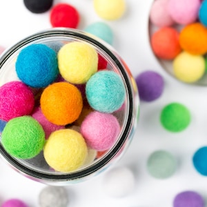 May include: A glass jar filled with colourful felt balls. The jar is on a white surface with more felt balls scattered around it.