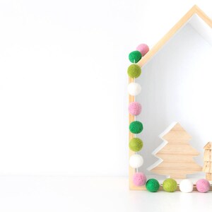 May include: A wooden house-shaped shelf with a garland of pink, white, and green felt balls. The shelf has a cutout of a tree and two smaller trees inside.