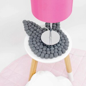 May include: A pink lamp with a chrome base sits on a white stool with a gray felt ball bunny-shaped coaster. The stool has wooden legs.