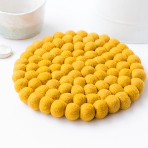 May include: A circular, yellow felt trivet composed of numerous small felt balls. The trivet is a vibrant, mustard yellow hue and rests on a white surface. A white ceramic mug and spoons are in the background.
