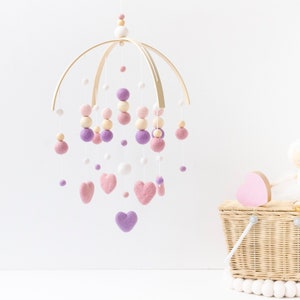 May include: A wooden baby mobile with pink, purple, and white felt balls and hearts hanging from a circular wooden frame. The mobile is hanging from the ceiling.