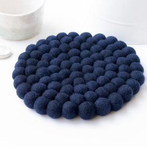 May include: A navy blue felt ball trivet, circular in form, featuring a close arrangement of small, spherical felt balls. The trivet is designed to protect surfaces from heat. White ceramic mug and spoons are in the background.