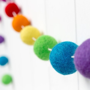 May include: A garland featuring a series of spherical felt balls in a vibrant rainbow color sequence, including red, orange, yellow, green, blue, and purple. Each individual felt ball is strung onto a thin white cord, creating a continuous line.