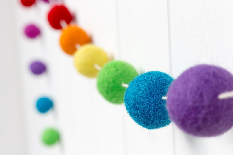May include: A garland featuring a series of spherical felt balls in a vibrant rainbow color sequence, including red, orange, yellow, green, blue, and purple. Each individual felt ball is strung onto a thin white cord, creating a continuous line.
