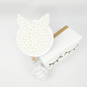 May include: White felt ball garland in the shape of a bunny head, on a white pedestal with a wooden base. A white knotted rope ball is in the foreground.
