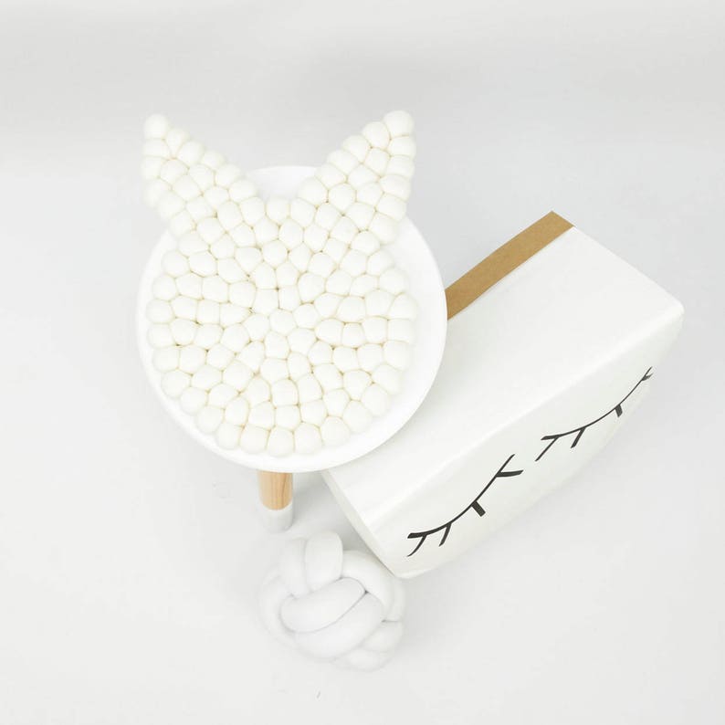 May include: White felt ball garland in the shape of a bunny head, on a white pedestal with a wooden base. A white knotted rope ball is in the foreground.