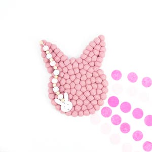 May include: A pink felt bunny wall hanging with a white wooden bunny charm and a white and natural wood bead garland.