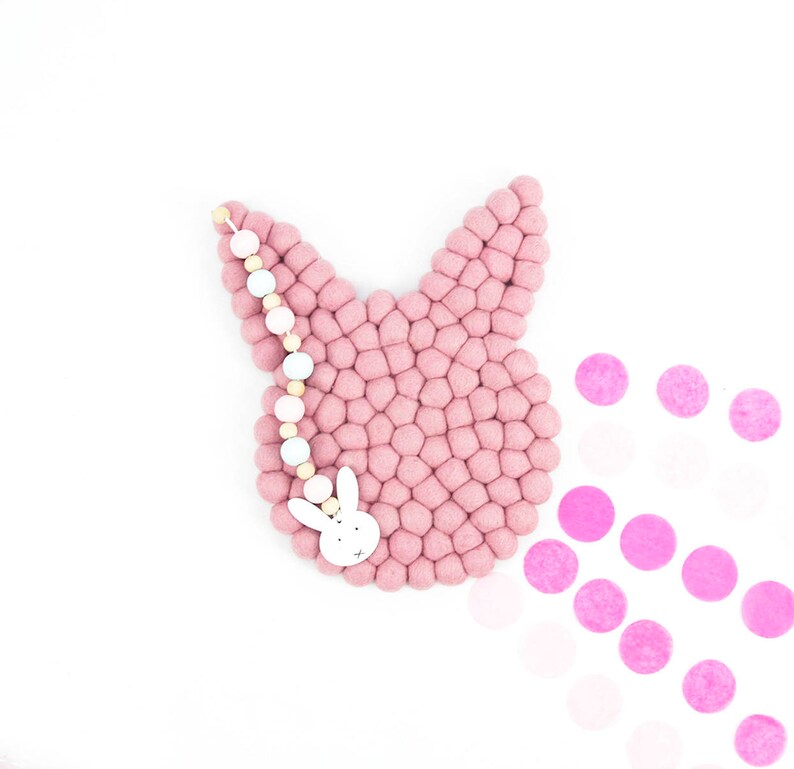 May include: A pink felt bunny wall hanging with a white wooden bunny charm and a white and natural wood bead garland.