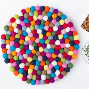 May include: A round, colorful felt ball rug with a mosaic pattern. The rug is composed of various colored felt balls, including red, blue, green, yellow, and purple. A white square bowl of almonds and a small glass bowl with rosemary sprigs are also in the image.