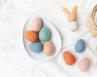 May include: A white plate with six felt Easter eggs in various colors, including blue, green, orange, and pink. The plate is on a white fabric background with two wooden bunny figurines.