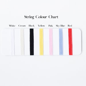 May include: A String Colour Chart displaying seven vertical strands of thread in white, cream, black, yellow, pink, sky blue, and red. Each color is labeled above the corresponding thread on a white rectangular card for easy identification.