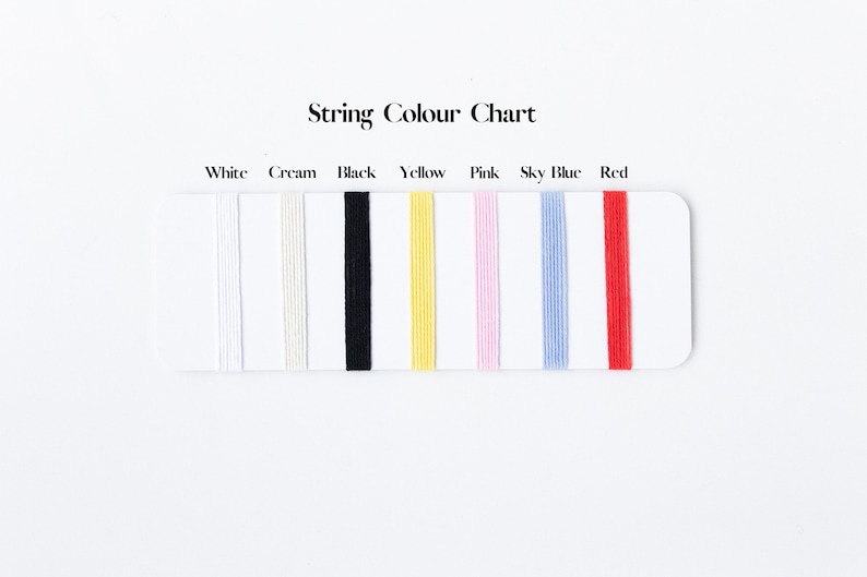 May include: A String Colour Chart displaying seven vertical strands of thread in white, cream, black, yellow, pink, sky blue, and red. Each color is labeled above the corresponding thread on a white rectangular card for easy identification.