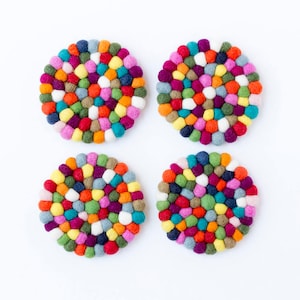 May include: Four colourful round felt coasters with a circular pattern of multi-coloured felt balls. The coasters are arranged in a square pattern on a white background.