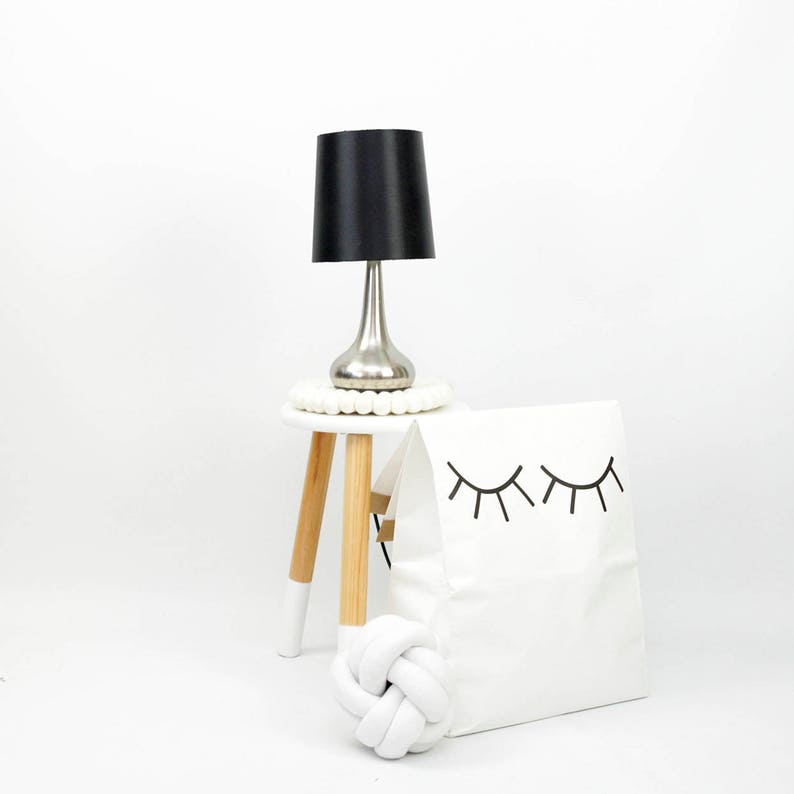 May include: A black table lamp with a silver base sits on a white stool with wooden legs. A white storage bag with black eyelashes printed on it is next to the stool. A white knotted pillow is in front of the bag.