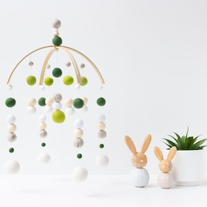 May include: A wooden baby mobile with green, white, and gray felt balls hanging from a wooden hoop. Two wooden bunny figurines are in the foreground.