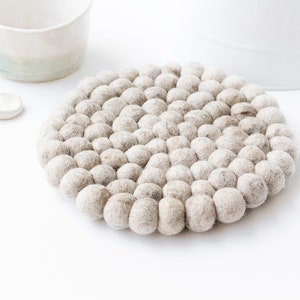 May include: A circular, pale grey felt trivet composed of numerous small, tightly-packed felt balls. The trivet sits on a white surface, with a white mug and two small spoons in the background. Designed to protect surfaces from heat.