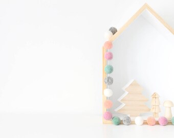 May include: A wooden house shaped shelf with a white interior. A garland of pastel colored felt balls hangs from the top of the shelf. There are three wooden ornaments on the shelf, including a tree and two mushrooms.