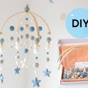 May include: A handmade baby mobile with a wooden frame, featuring felt balls and star shapes in shades of blue, gray, and cream. The mobile is suspended, with a DIY kit in a box nearby. The text "DIY" is in a blue circle.