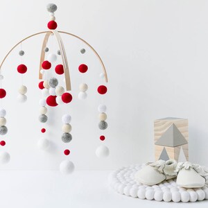 May include: A wooden baby mobile with red, white, and gray felt balls hanging from a wooden hoop. The mobile is hanging over a white surface. There is a white rug with white pom-poms on the floor and a wooden block with a gray triangle on top of it.