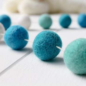 May include: A garland of white, teal, and blue felt balls strung together with white thread. The garland is on a white wooden surface.