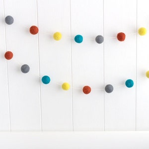 May include: A garland of felt balls in grey, teal, yellow, and orange. The balls are strung on a white string and are hanging from a white wall.