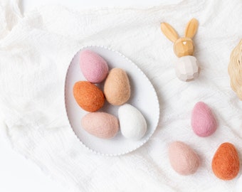 May include: A white plate with six felt Easter eggs in various shades of pink, orange, and white. The eggs are arranged in a circle on the plate. There is a wooden bunny figurine and a wicker basket with a felt egg in the background.